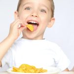 A child with pleasure eating potato chips sitting at a table on a white background. The boy is eating chips and smiling. 4k