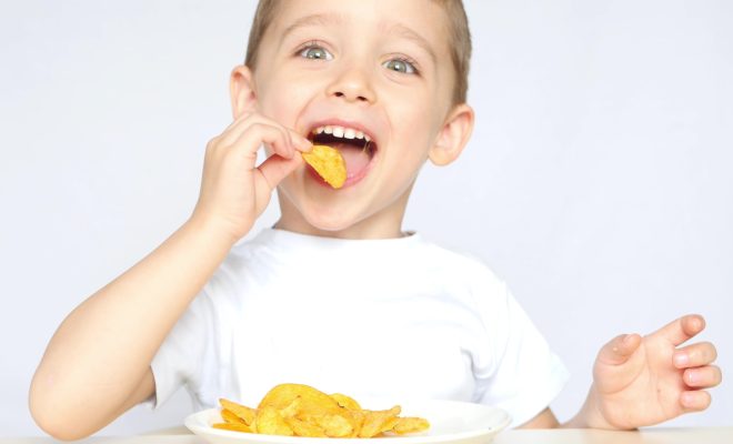 A child with pleasure eating potato chips sitting at a table on a white background. The boy is eating chips and smiling. 4k