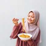 Portrait of Young Muslim Woman in Hijab Enjoying Spicy Instant Noodles Indoors with Fork and Bowl in Hand