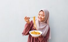 Portrait of Young Muslim Woman in Hijab Enjoying Spicy Instant Noodles Indoors with Fork and Bowl in Hand