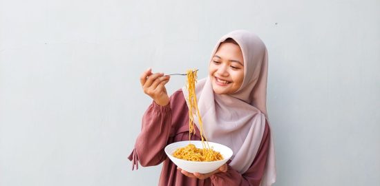 Portrait of Young Muslim Woman in Hijab Enjoying Spicy Instant Noodles Indoors with Fork and Bowl in Hand