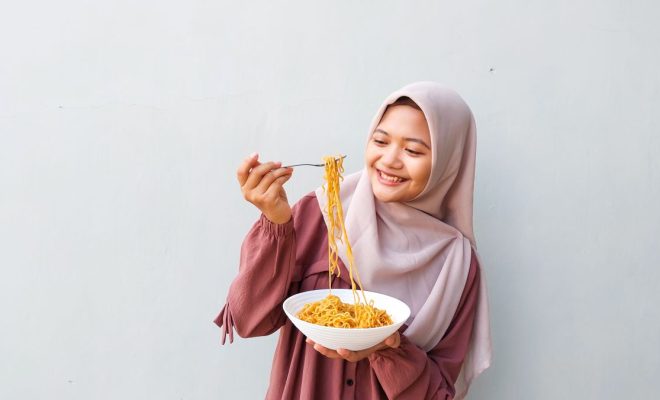 Portrait of Young Muslim Woman in Hijab Enjoying Spicy Instant Noodles Indoors with Fork and Bowl in Hand